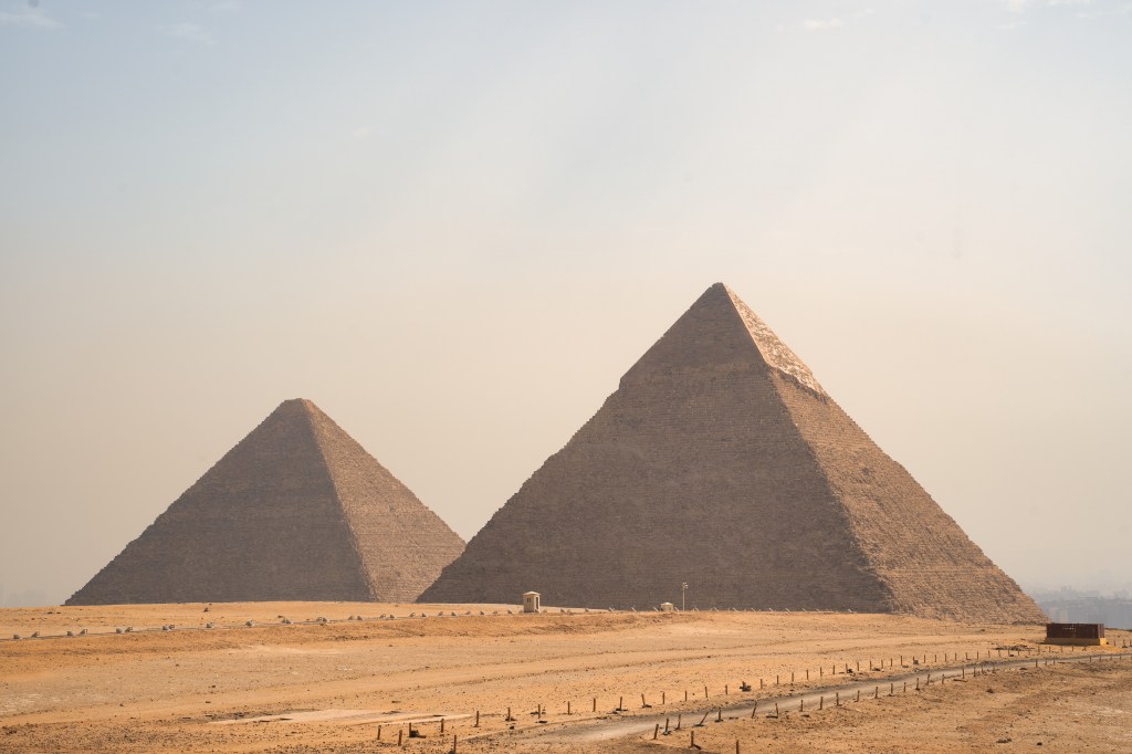 Two pyramids silhouetted against a pale sky across the sandy Giza plateau.