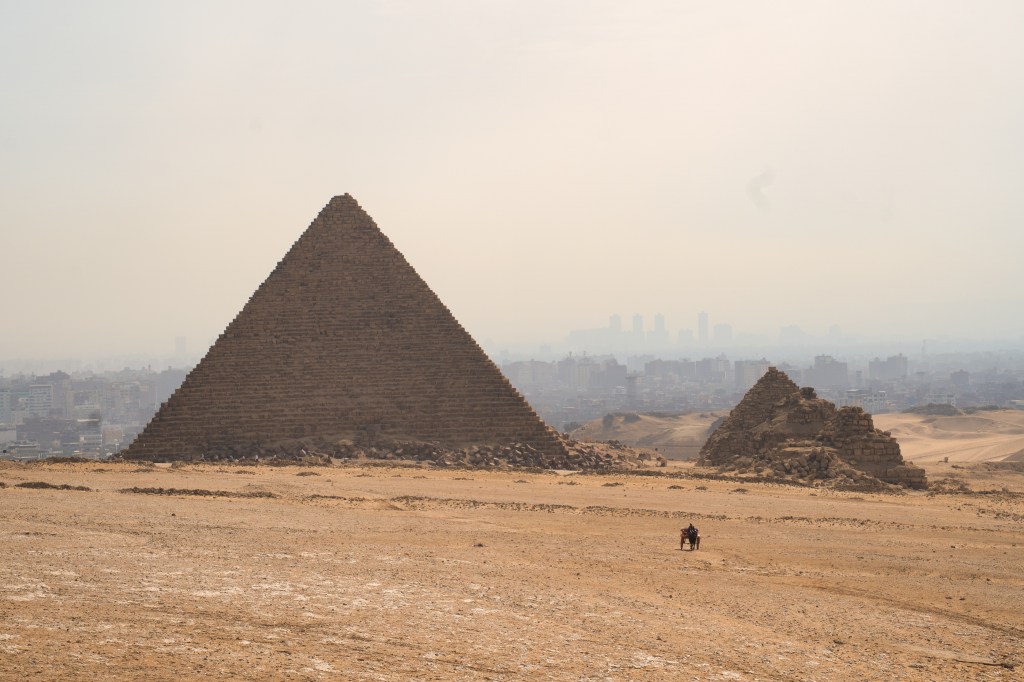 A pyramid and smaller ruins on the sandy plateau, with the city beyond.