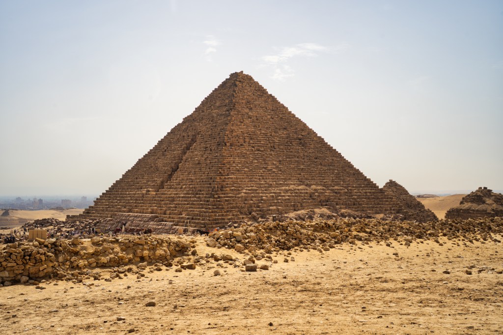 A pyramid rising beyond low stone ruins and sandy ground.