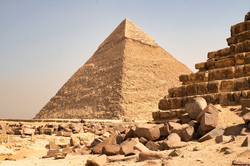 A pyramid with pale stone near its summit rising behind broken blocks and ancient walls.