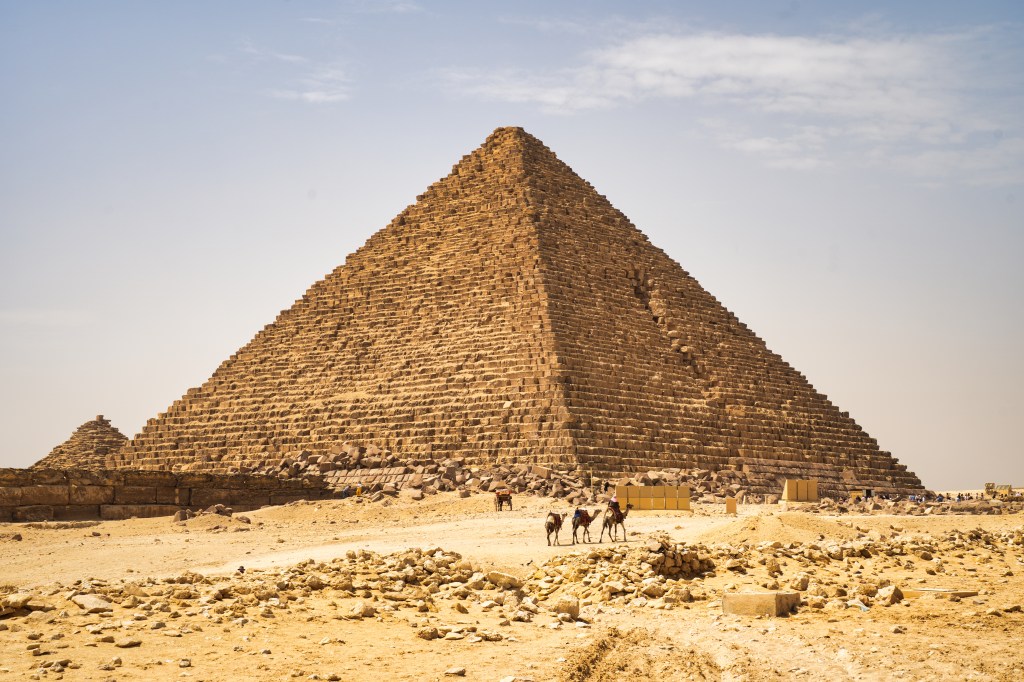 Camel riders crossing sandy ground in front of a weathered pyramid.