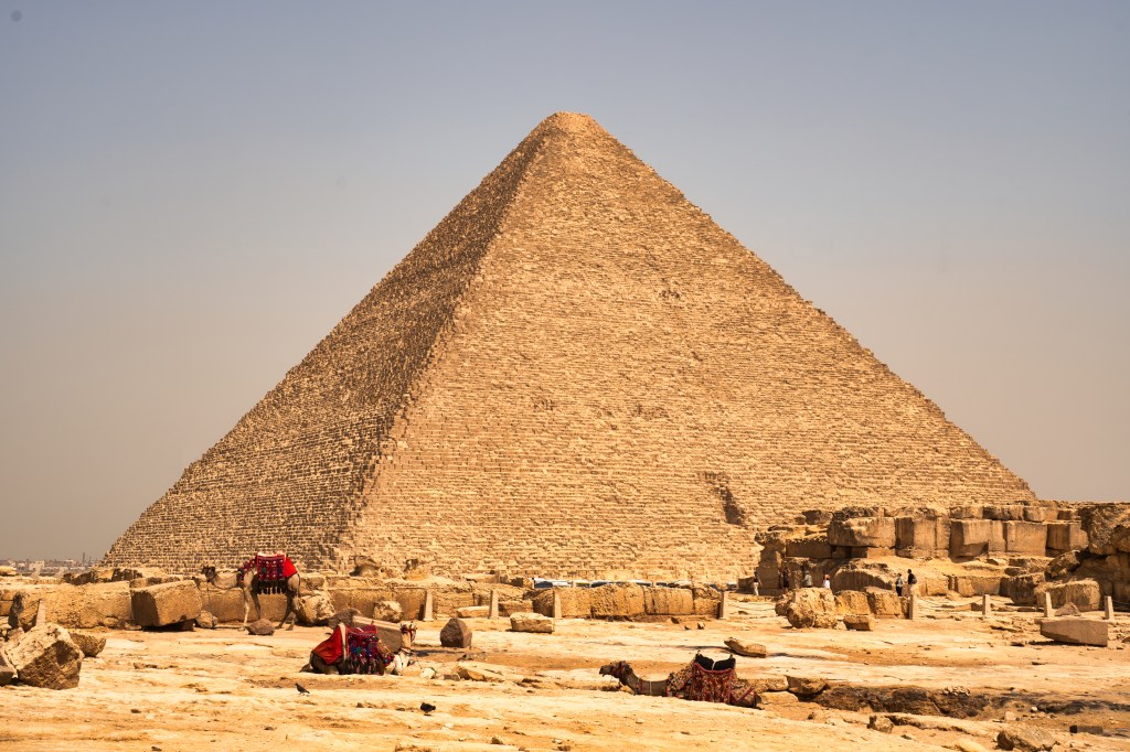 A sunlit pyramid above scattered stone blocks and small groups of visitors.