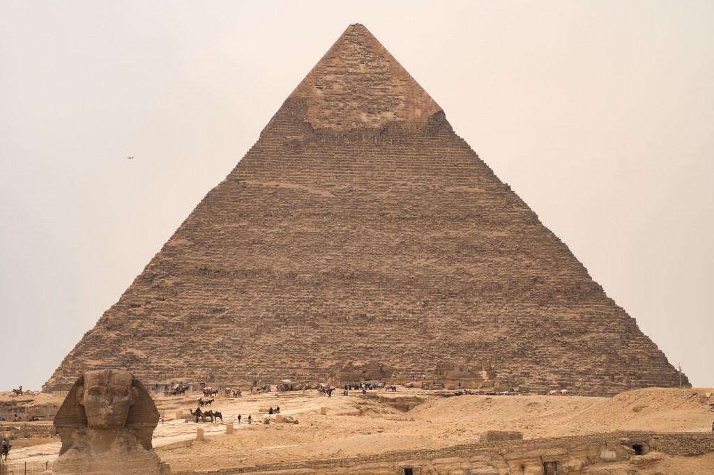 A tall pyramid with a smooth cap above the Sphinx and distant visitors.