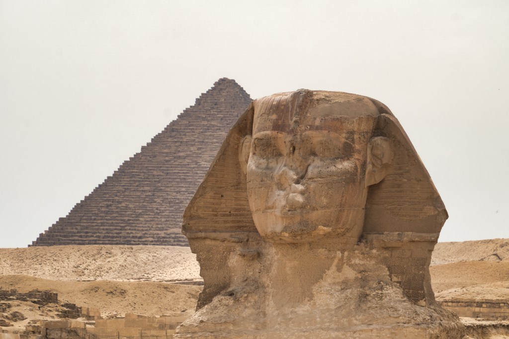 The weathered face of the Sphinx with a pyramid behind it.