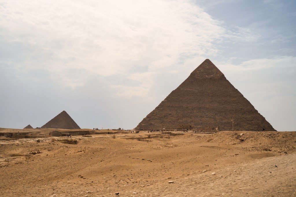 Two pyramids beyond a broad stretch of sandy desert under a lightly clouded sky.