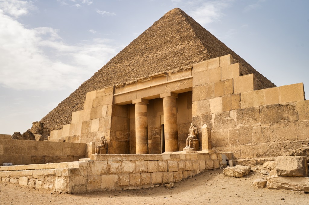 A colonnaded stone entrance and weathered walls with a pyramid rising behind them.