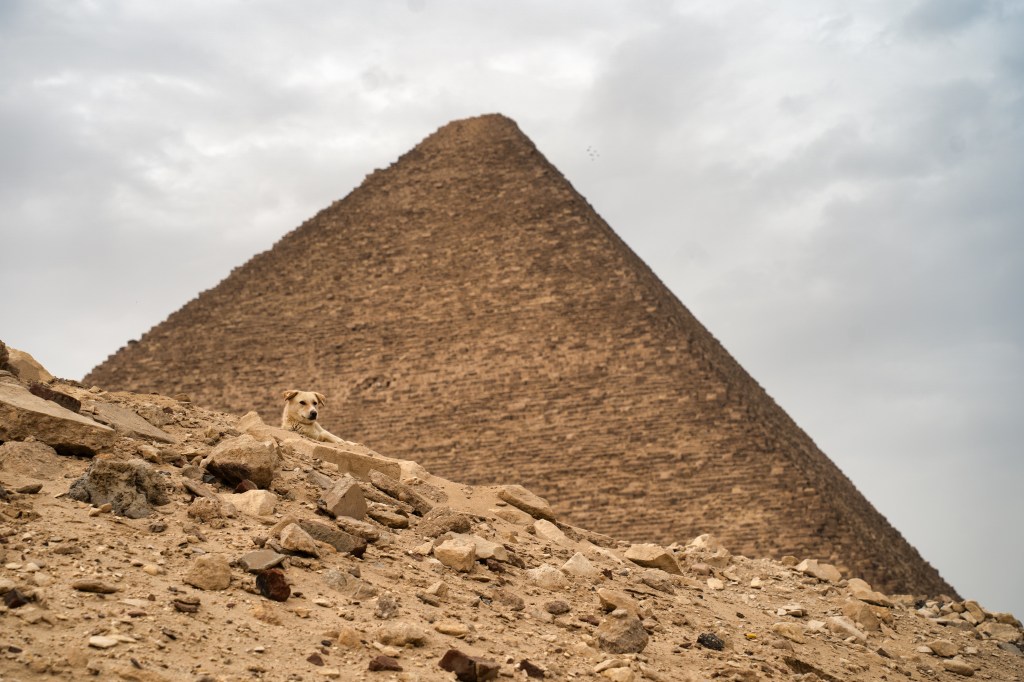 A pyramid rising behind broken stone and rocky ground under grey clouds.