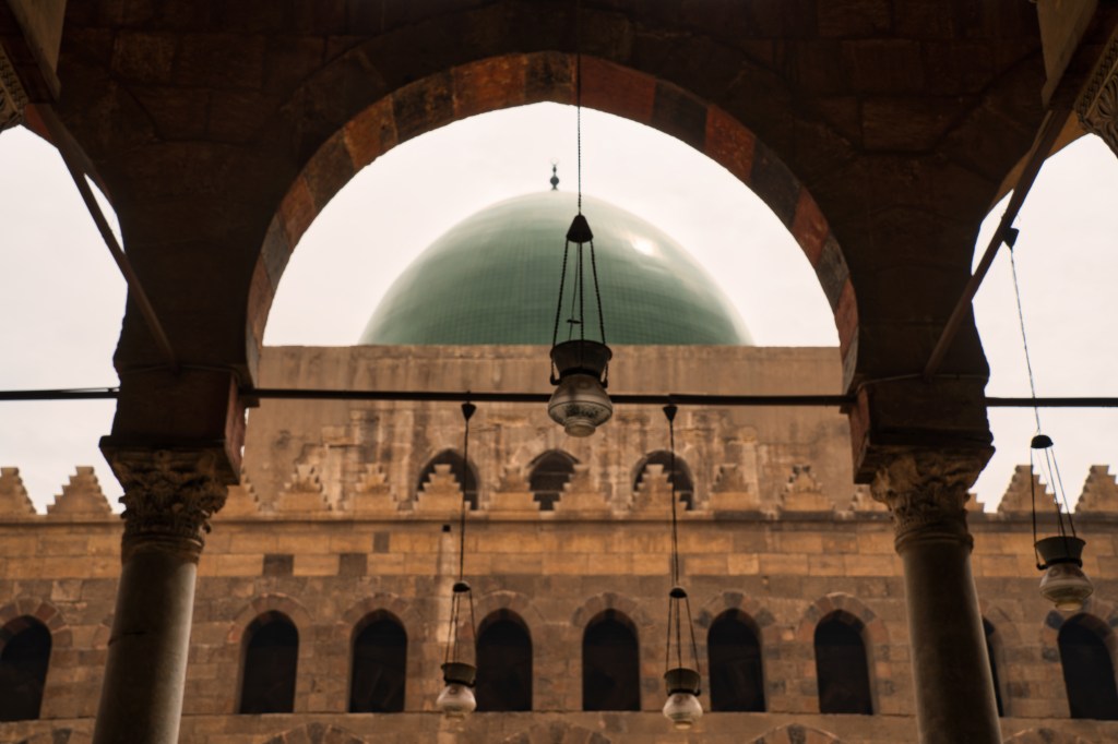 A green mosque dome seen through a shaded courtyard arch.