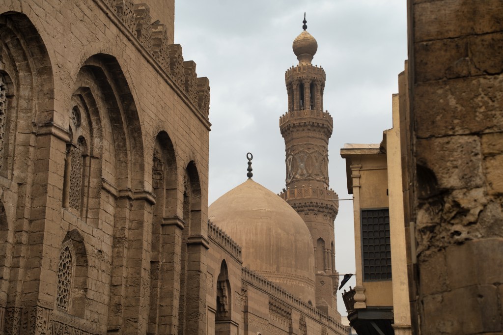 A mosque dome and minaret framed by old stone buildings.