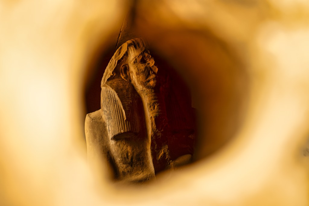 A seated stone statue glimpsed through a small opening in a wall.