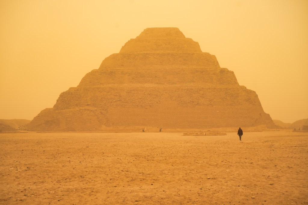 A stepped pyramid in golden haze, with a lone figure in the sandy foreground.