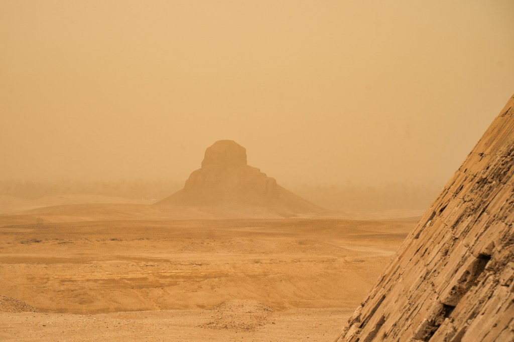 A distant monument in desert haze, seen beside the sloping stones of a pyramid.