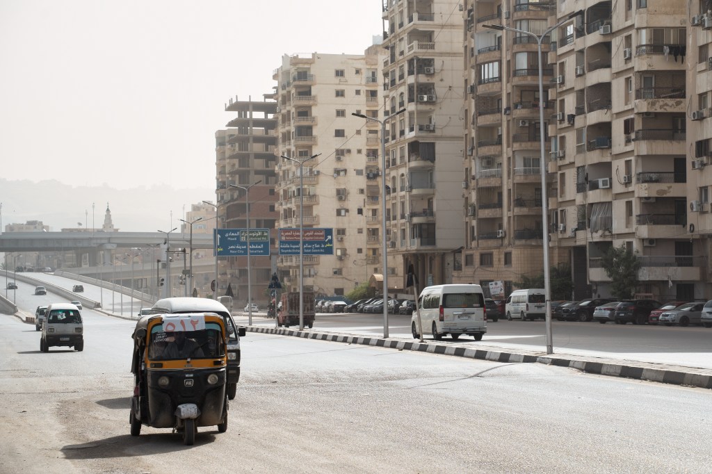 A tuk-tuk travelling along a broad road lined with apartment buildings.